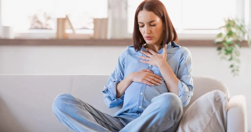Pregnant woman sitting on a couch, hands on her stomach, visibly experiencing breathing problems in pregnancy.