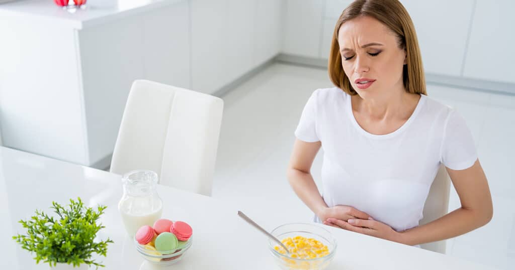 A woman sits at a table with a bowl of cereal, clutching her stomach and wearing a pained expression, showing symptoms of food poisoning and stomach flu.
