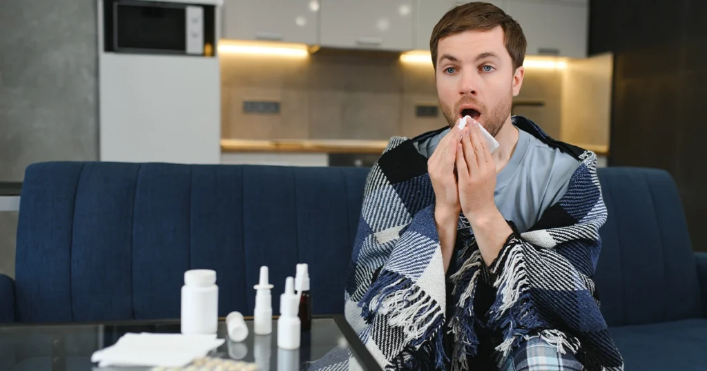 Sick man coughing on couch with tissues and medicine