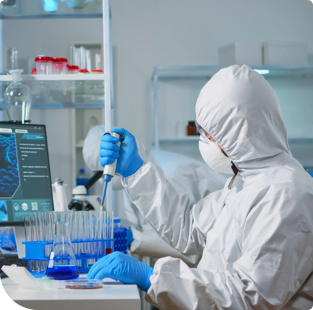 A scientist in a full hazmat suit and blue gloves using a pipette to transfer liquids into test tubes in a medical research laboratory.