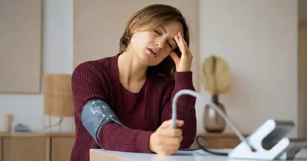 Woman checking blood pressure at home while holding her head in discomfort.