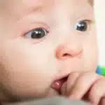Baby sucking thumb, possibly showing teething symptoms and runny nose.