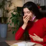A young woman in a red sweater sitting at a desk and coughing while holding her chest