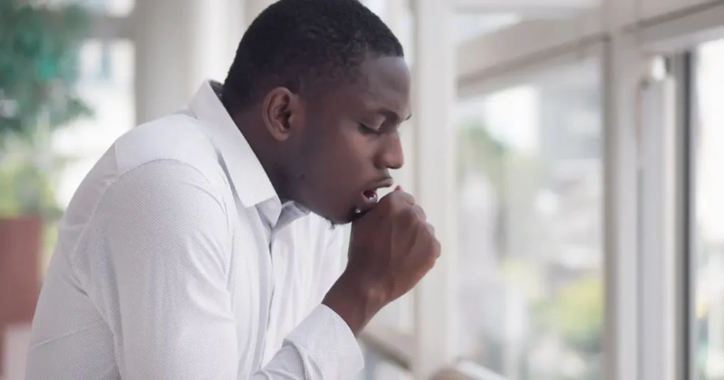 A professional stock photo of a young man in a white button-down shirt, coughing into his hand while standing near a large window in a bright office or clinic setting. The side-profile shot conveys physical discomfort and symptoms of a persistent cough.