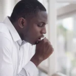 A professional stock photo of a young man in a white button-down shirt, coughing into his hand while standing near a large window in a bright office or clinic setting. The side-profile shot conveys physical discomfort and symptoms of a persistent cough.