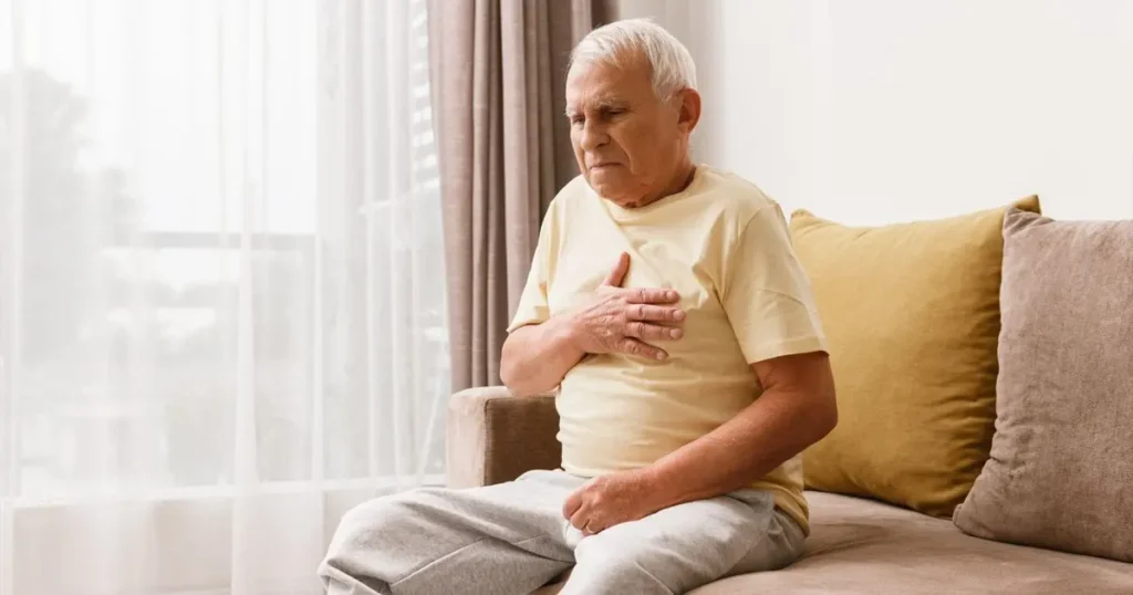 Elderly man sitting on a brown sofa in natural light, clutching his chest with a pained expression, illustrating chest pain or heart-related discomfort.