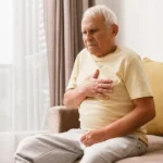 Elderly man sitting on a brown sofa in natural light, clutching his chest with a pained expression, illustrating chest pain or heart-related discomfort.
