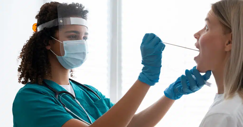 A healthcare worker in scrubs and a face shield uses a swab to perform a medical test on a patient’s throat.