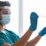 A healthcare worker in scrubs and a face shield uses a swab to perform a medical test on a patient’s throat.