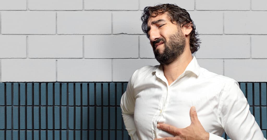 A man in a white shirt winces in pain while clutching his chest and stomach, illustrating symptoms of acid reflux.
