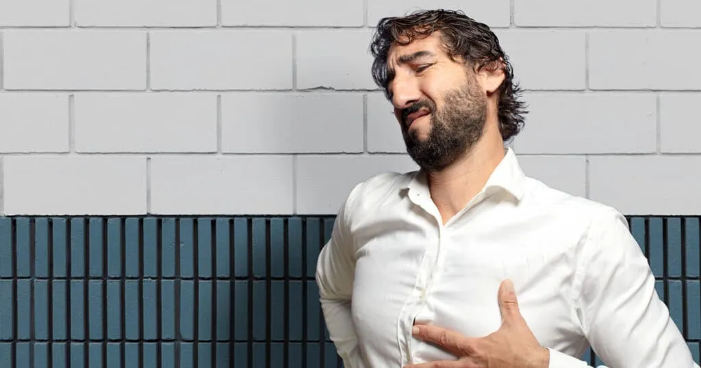A man in a white shirt winces in pain while clutching his chest and stomach, illustrating symptoms of acid reflux.