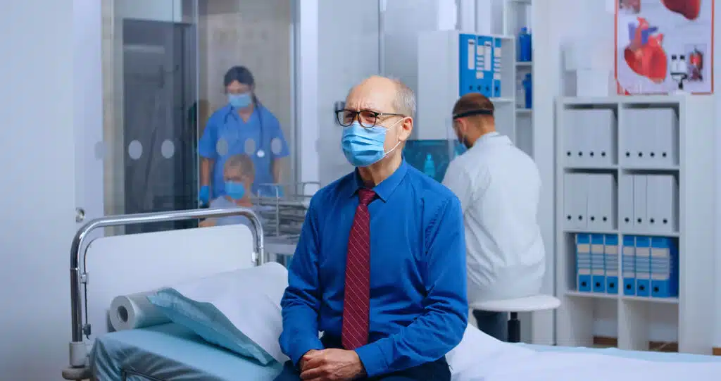 An elderly man wearing a face mask and tie sits on a hospital bed while medical staff work in the background.