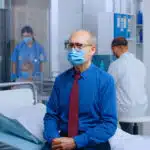 An elderly man wearing a face mask and tie sits on a hospital bed while medical staff work in the background.
