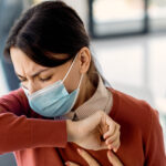 Masked woman coughing into her elbow inside an office setting.
