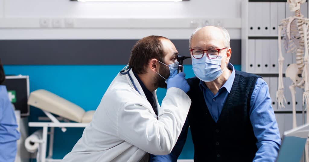 A doctor in a white lab coat uses an otoscope to examine the ear of an elderly man wearing a surgical mask in a medical office.