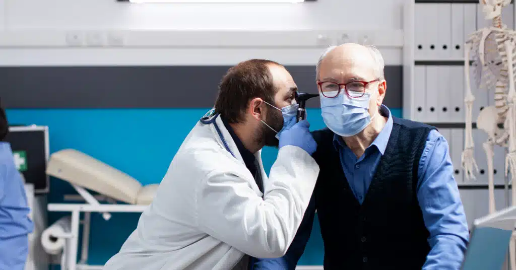A doctor in a white lab coat uses an otoscope to examine the ear of an elderly man wearing a surgical mask in a medical office.