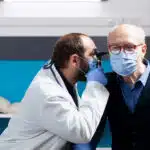 A doctor in a white lab coat uses an otoscope to examine the ear of an elderly man wearing a surgical mask in a medical office.