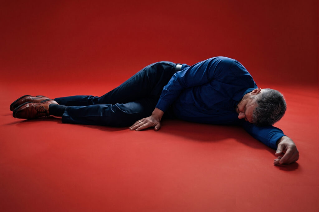 A man wearing a blue button-down shirt, dark trousers, and brown dress shoes lying flat on a red studio surface.