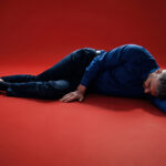 A man wearing a blue button-down shirt, dark trousers, and brown dress shoes lying flat on a red studio surface.
