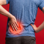 A person viewed from behind touching their lower back, with a bright red, glowing spot highlighting lower back pain against a solid red background.