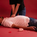A person performing CPR on an unconscious man lying on the floor near a bottle of pills.