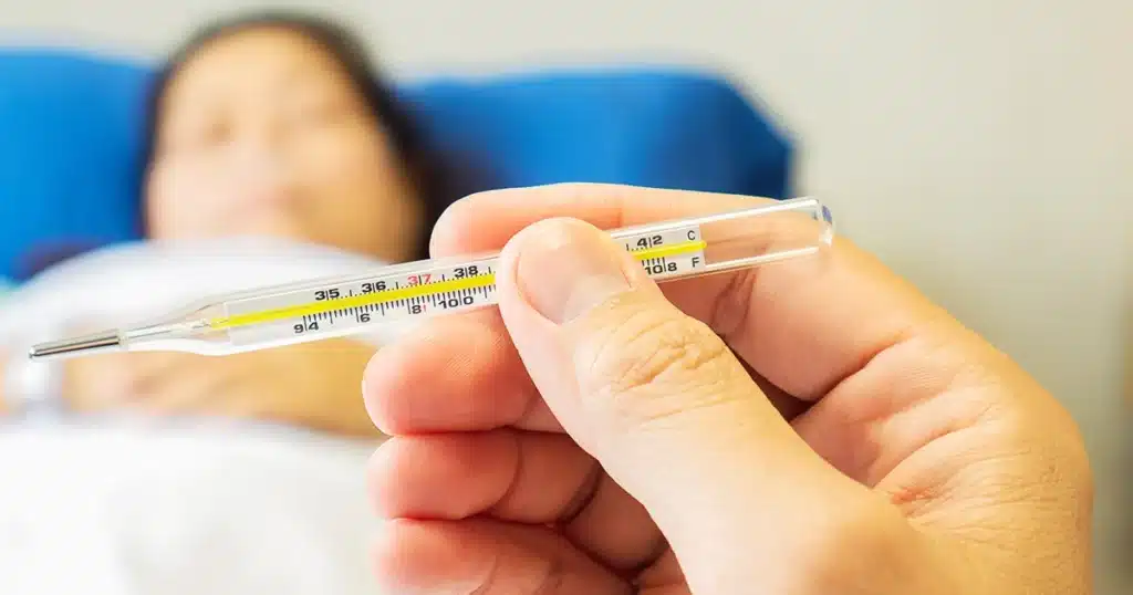 A hand holding a thermometer with a high reading while a sick person rests in bed.