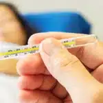 A hand holding a thermometer with a high reading while a sick person rests in bed.