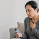 Adult in their 30s experiencing asthma symptoms while seated near a window.