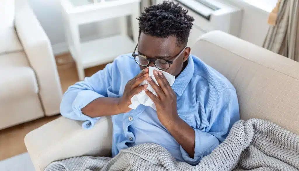 Man sitting on sofa blowing nose with tissue while sick.