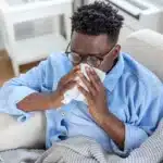 Man sitting on sofa blowing nose with tissue while sick.