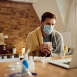 Sick man wearing a face mask and wrapped in a blanket while working on a laptop.
