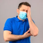 Man wearing a blue face mask holding his hand to his forehead with a headache.