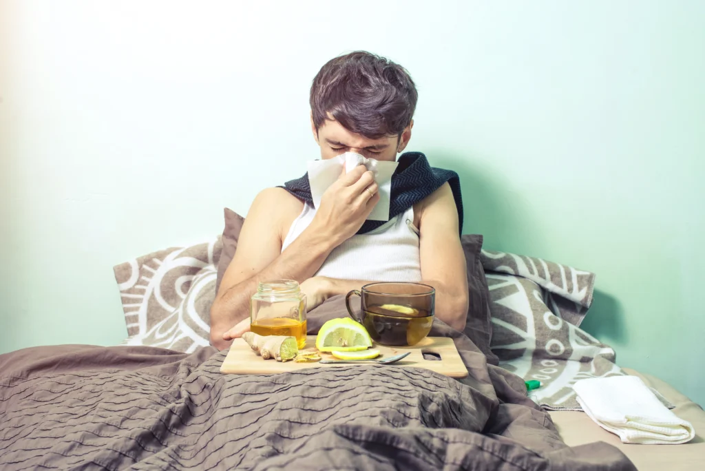 Sick man blowing his nose in bed next to a tray with lemon, ginger, and honey tea.