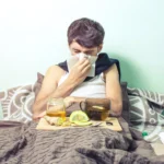 Sick man blowing his nose in bed next to a tray with lemon, ginger, and honey tea.