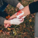 Person applying white gauze bandage to bloody arm in grass.